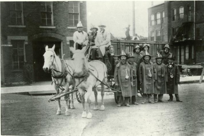 Historic photo of firefighters standing in front of and sitting on a horse-driven hose cart. 